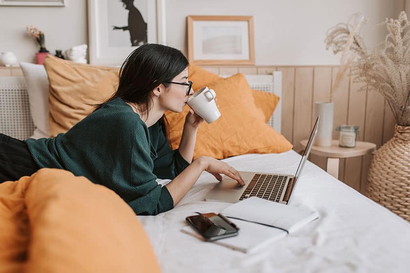 woman lying in bed drinking coffee and using laptop