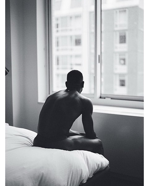 black and white portrait of a black man sitting on the edge of a bed looking out the window, with his back to the camera