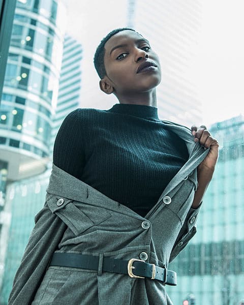 street portrait of a black person with close-cropped hair wearing a black turtleneck and grey belted trench coat