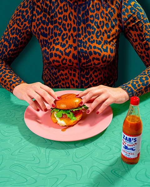 a figure wearing leopard print seated at a retro patterned table with a pink plate holding a sandwich in a bun, a bottle of hot sauce nearby