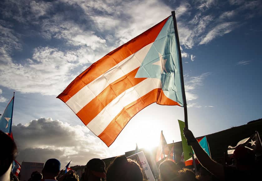 the flag of Puerto Rico held high above a crowd and lit from behind by the sun