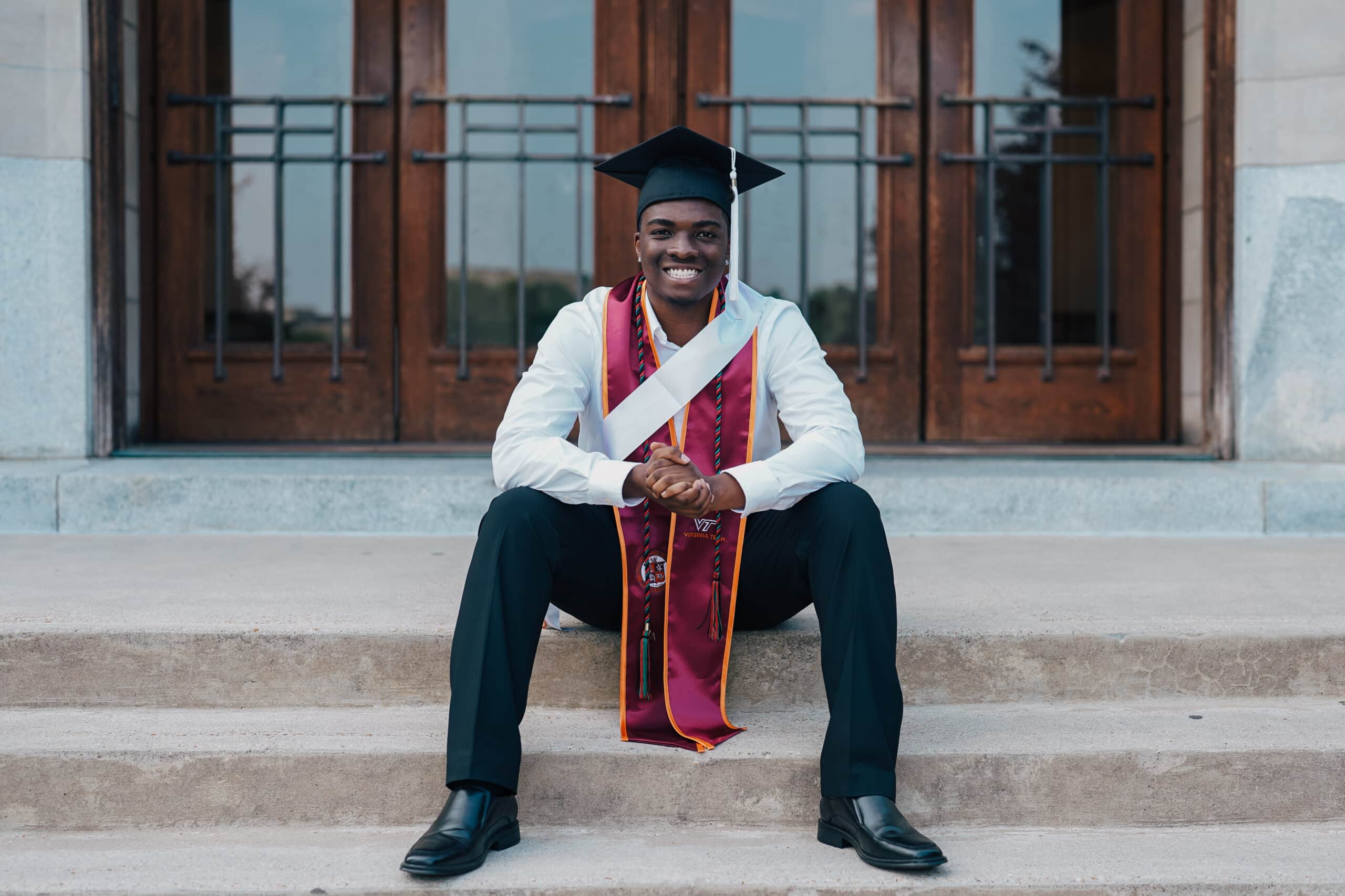 Senior portrait of African American boy sitting on steps wearing graduation cap and academic sash