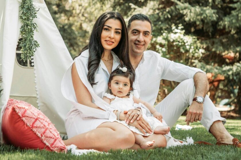 outdoor family portrait with mom dad and baby girl sitting on grass beside a tent and wearing white clothing