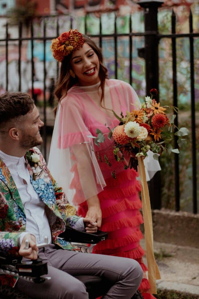 wedding portrait of colorful bride wearing red layered dress and groom in wheelchair wearing multicolor blazer strolling beside each other in a street