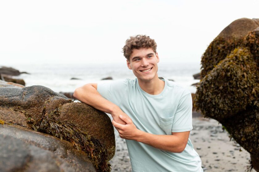 teen boy wearing tee shirt resting his elbow and leaning on a large rock by the ocean