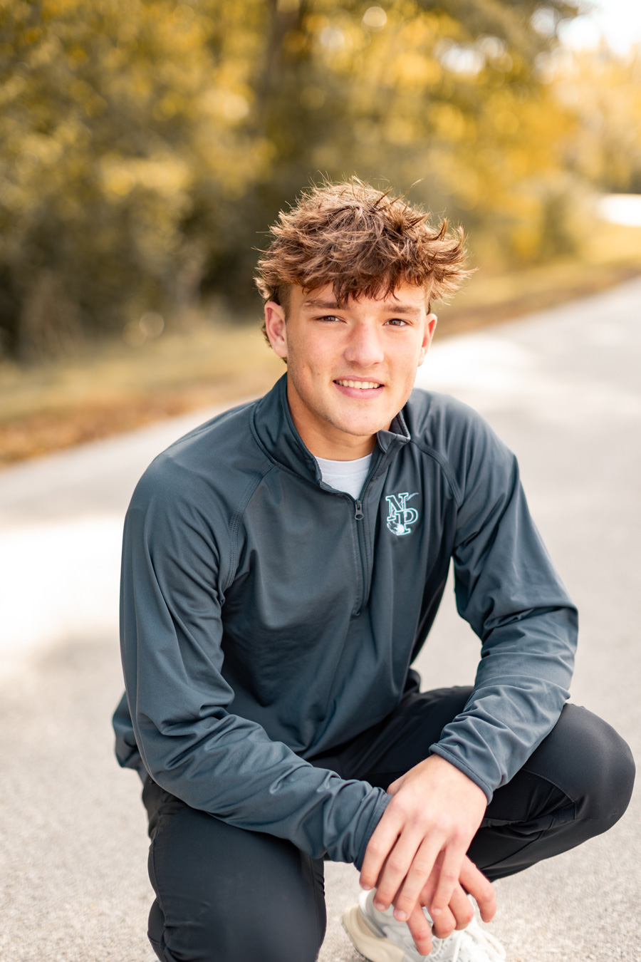 A smiling young man with messy brown hair crouches on a paved path, wearing a dark grey quarter-zip pullover and black pants against a blurred background of autumn trees.