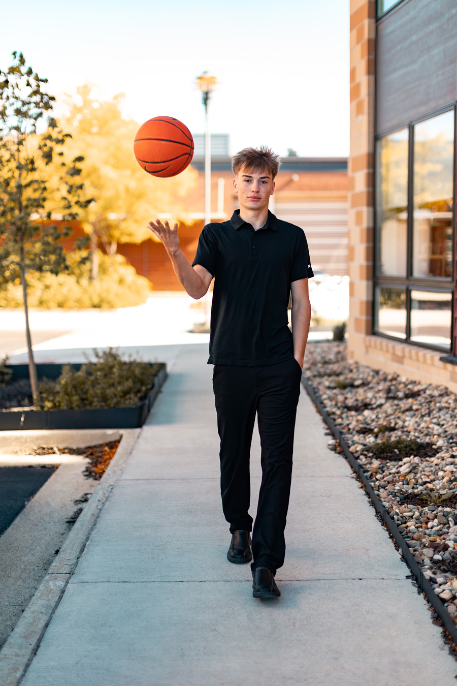 A young man dressed in all black walks forward on an outdoor concrete path while tossing a basketball in the air with his right hand.