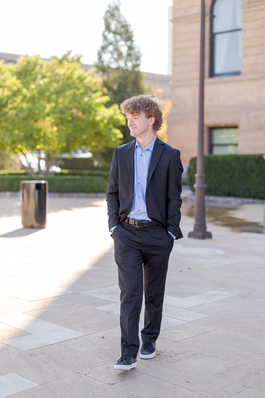 A young man with curly blonde hair smiles as he walks outdoors in a black suit and light blue button-down shirt with his hands in his pockets.