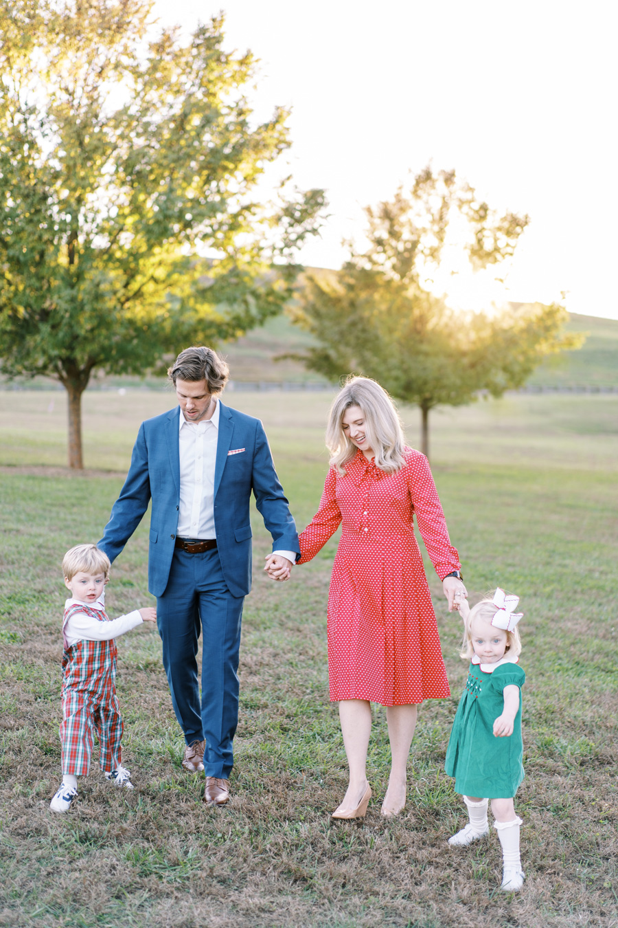 Family portrait: parents in formal wear holding hands with two young children in festive outfits, walking on a grassy field