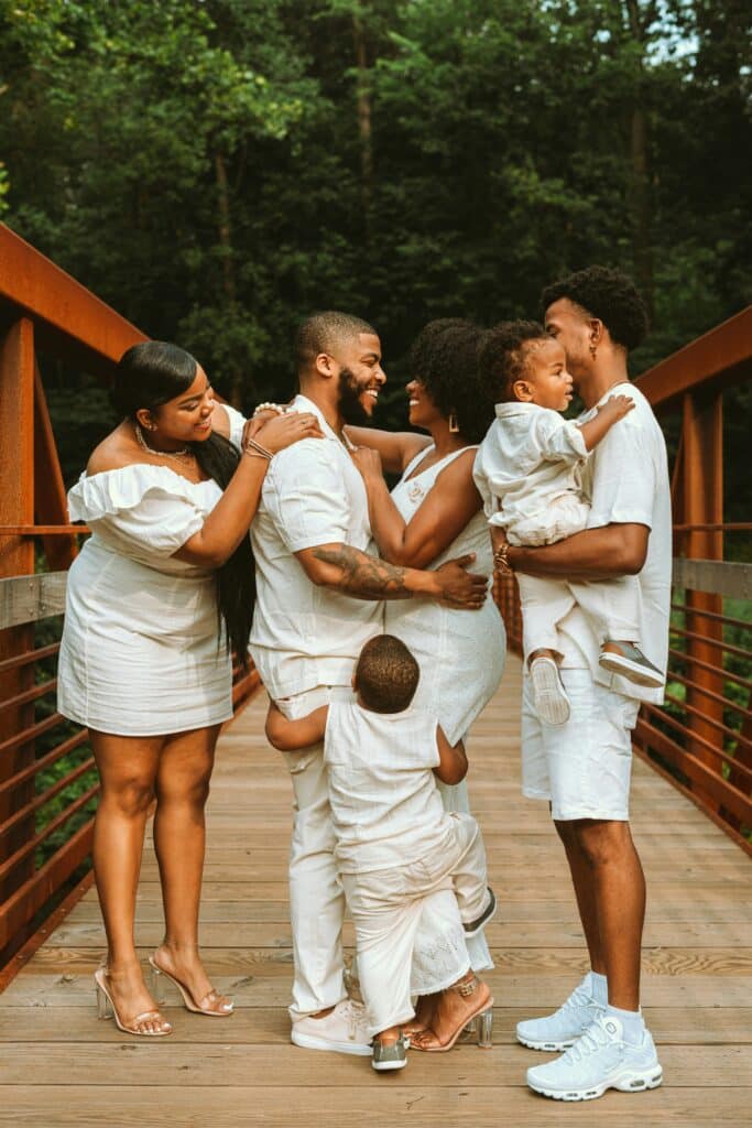 parents facing each other and smiling while their toddler hugs their legs, flanked by two teen children, one holding a toddler sibling.