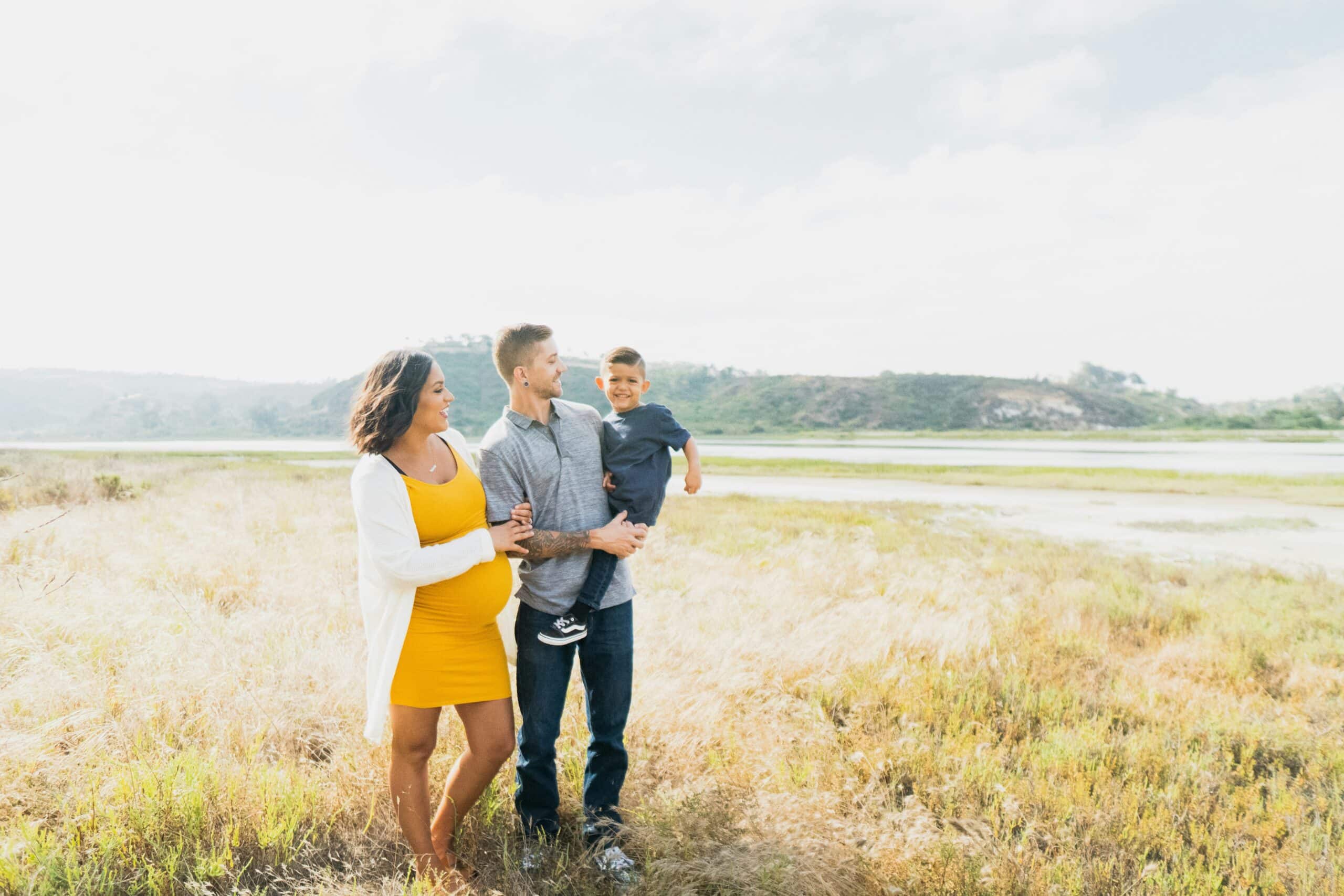 pregnant mom in yellow dress linking arms with husband holding toddler boy, standing together in large open field with distant mountains.