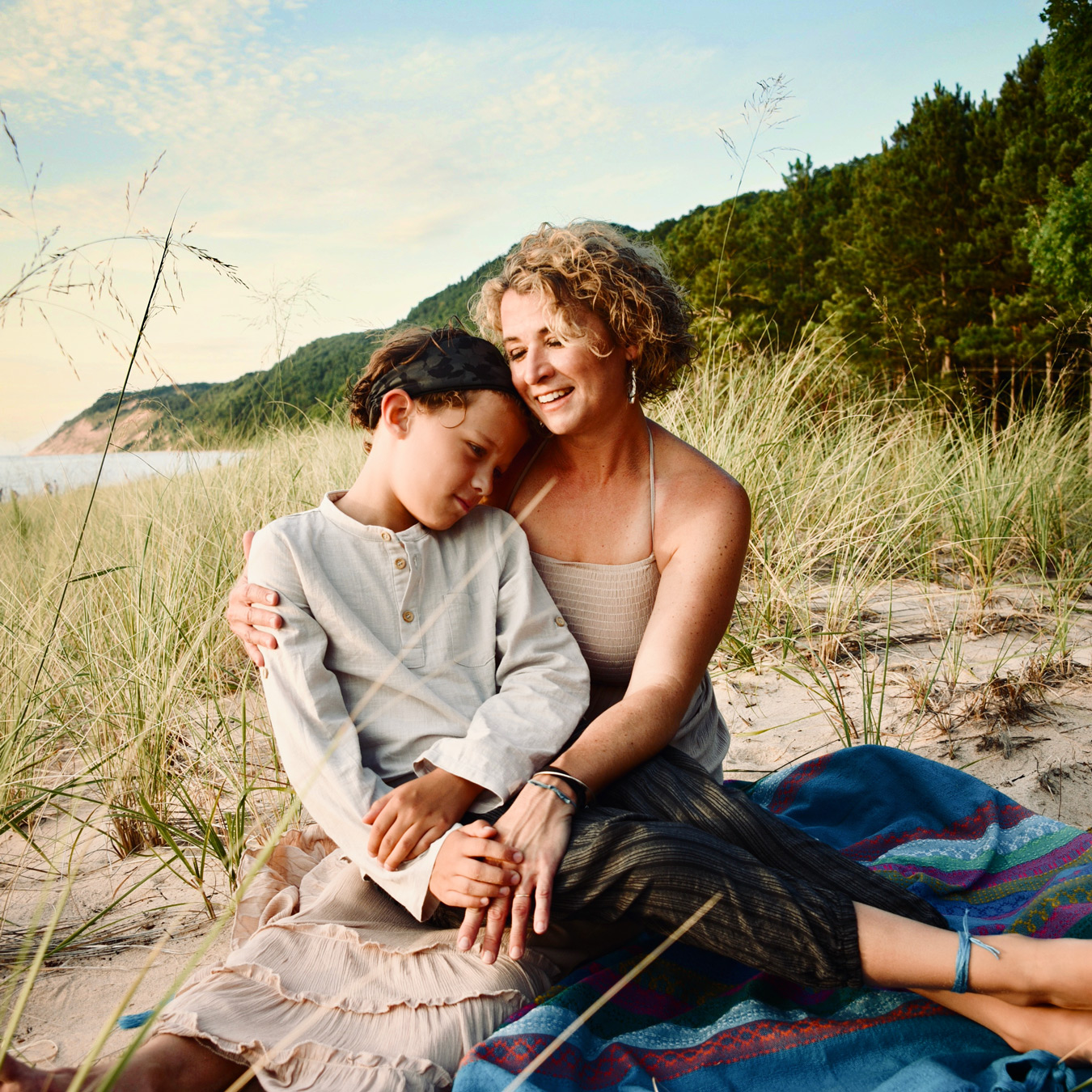 A woman with curly blonde hair sits on a colorful blanket on a sandy beach, embracing a young child with a dark headband who rests their head on her shoulder. Tall grasses surround them, with a forested hill and a glimpse of the ocean in the background under a partly cloudy sky. photographed by Noelle Biele