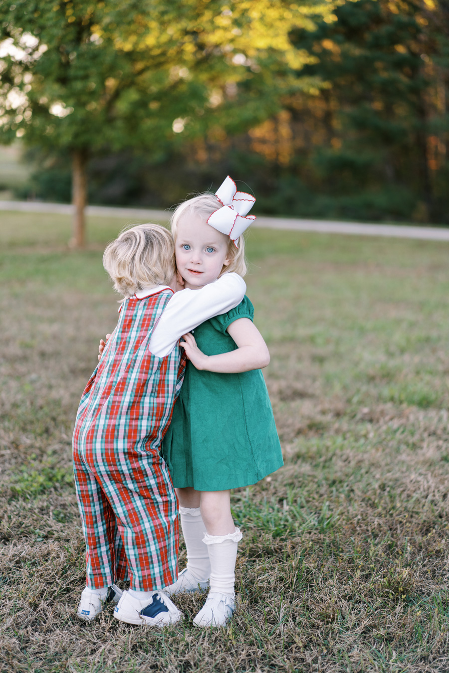 Two young children hugging outdoors. Girl in green dress and large white bow; boy in red and green plaid overalls