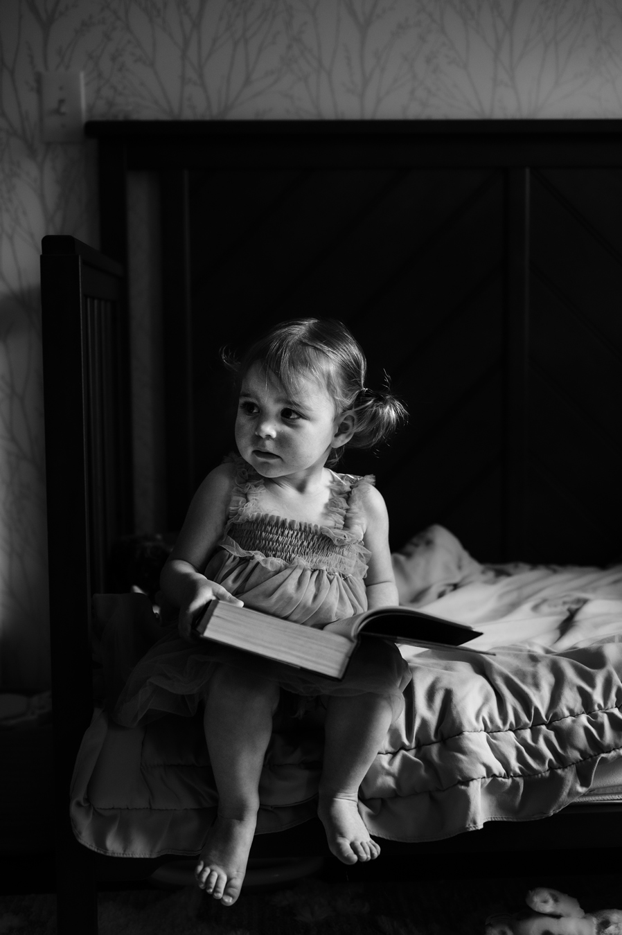 A black and white portrait shows a young girl with pigtails sitting on a bed, holding an open book and looking off to the side with a thoughtful expression. The girl is wearing a dress with ruffles, and the bed has a dark headboard and a textured comforter. photographed by Brooke Cadett