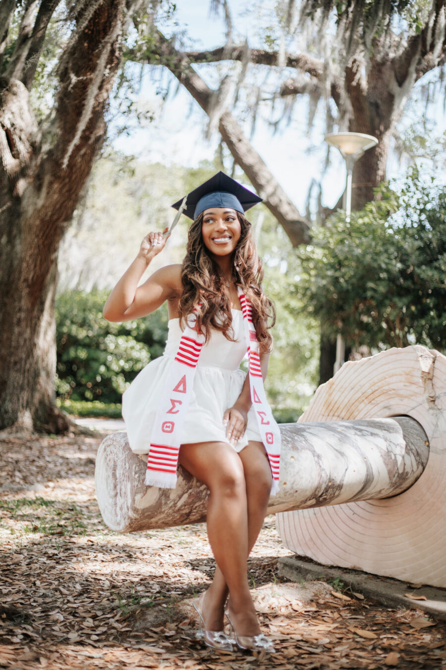 A smiling graduate in a white dress, graduation cap, and red-and-white Delta Sigma Theta stole sits on a large stone sculpture in a wooded park.