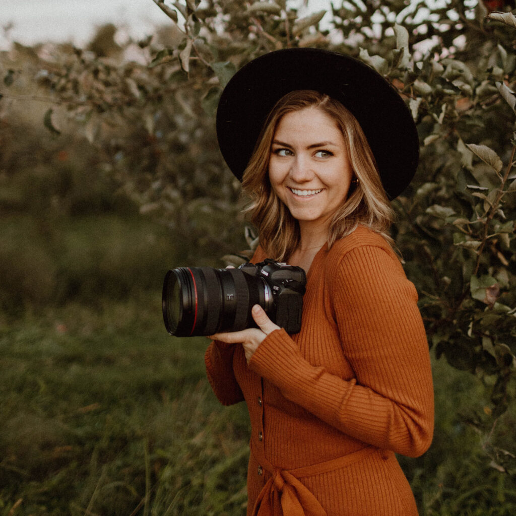Portrait of NC portrait photographer Heather Nelson standing beside an apple tree wearing a dark wide brim hat and orange sweater, holding her camera.