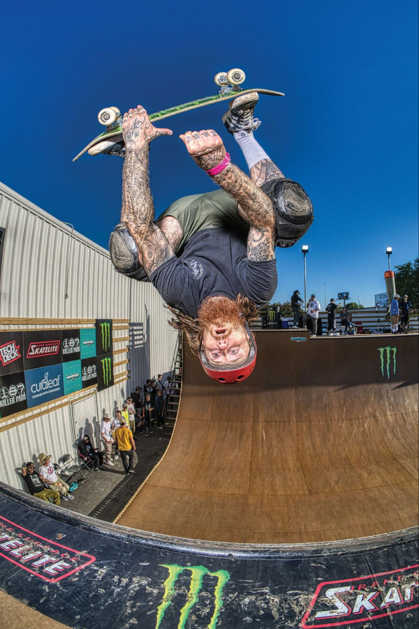 Tattooed skateboarder upside down, performing a trick in a ramp, spectators watching. Photo by Robert McClory.