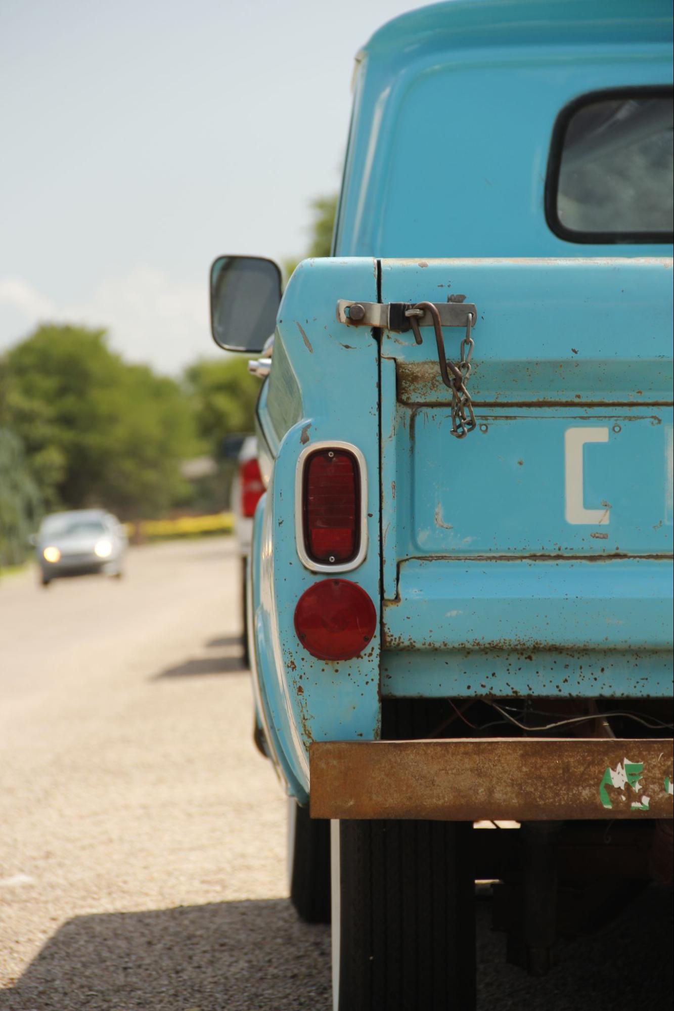 Close-up shot of the rear of a light blue vintage pickup truck, showing the tailgate with a chain latch, a partially visible license plate with the letter "C", and a rusty bumper. The truck is parked on a road with other vehicles visible in the background. Photo by Robert Smith.