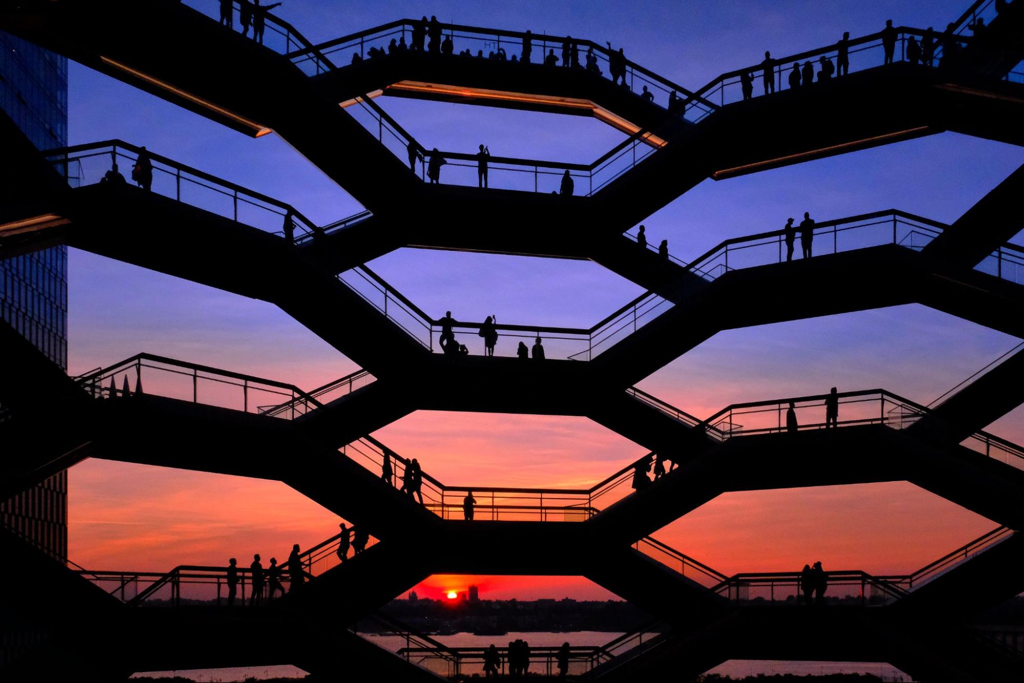 Silhouetted figures populate the interconnected stairways of the Vessel structure, framed against a vibrant sunset sky. The honeycomb-like design creates geometric patterns, with the setting sun visible through the lower openings. Photo by Tom Carroll.