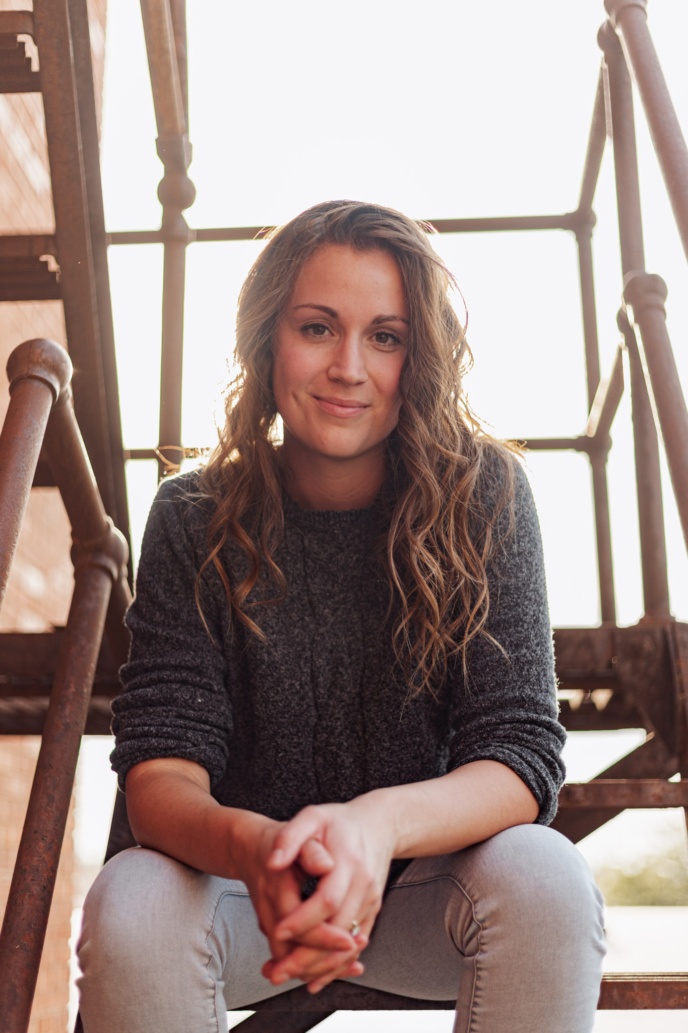 Woman with long brown hair, wearing a gray sweater and light jeans, sits on rusty metal stairs