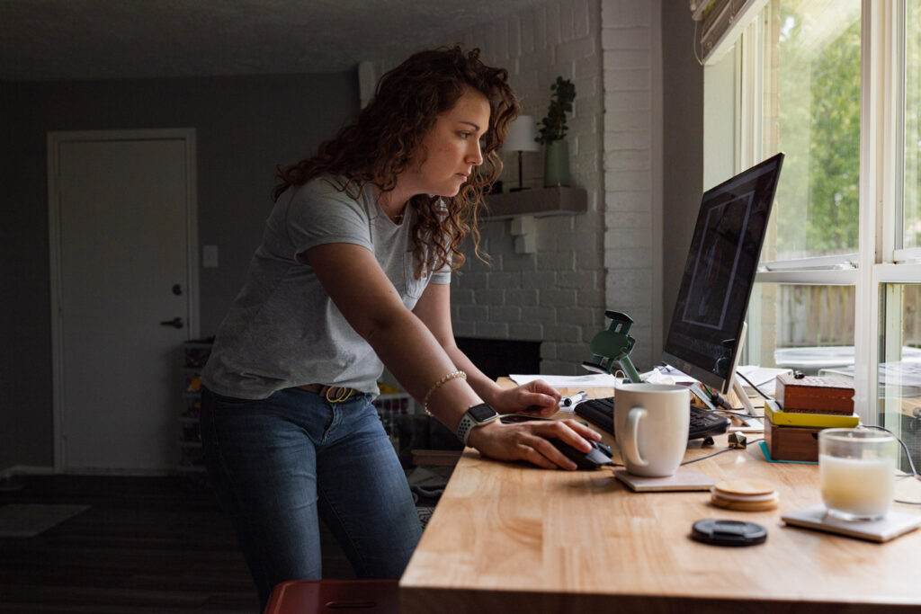 Woman with curly hair uses computer mouse at a light wood desk near a window. portrait of Caroline Stubbs