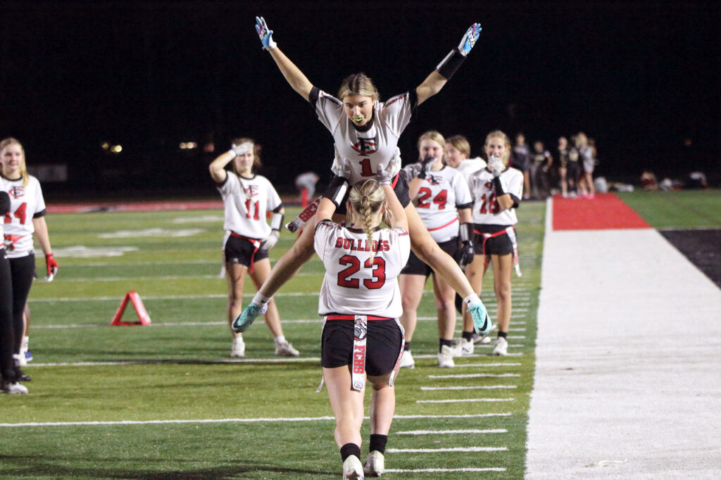 Female flag football player hoisted on teammate's shoulders, arms raised in victory; other players in background