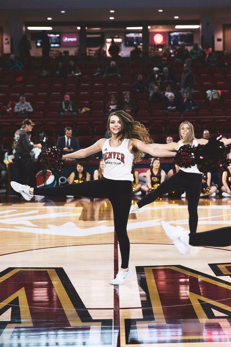 University of Denver dance team member performing a leg kick on the basketball court, holding pom-poms