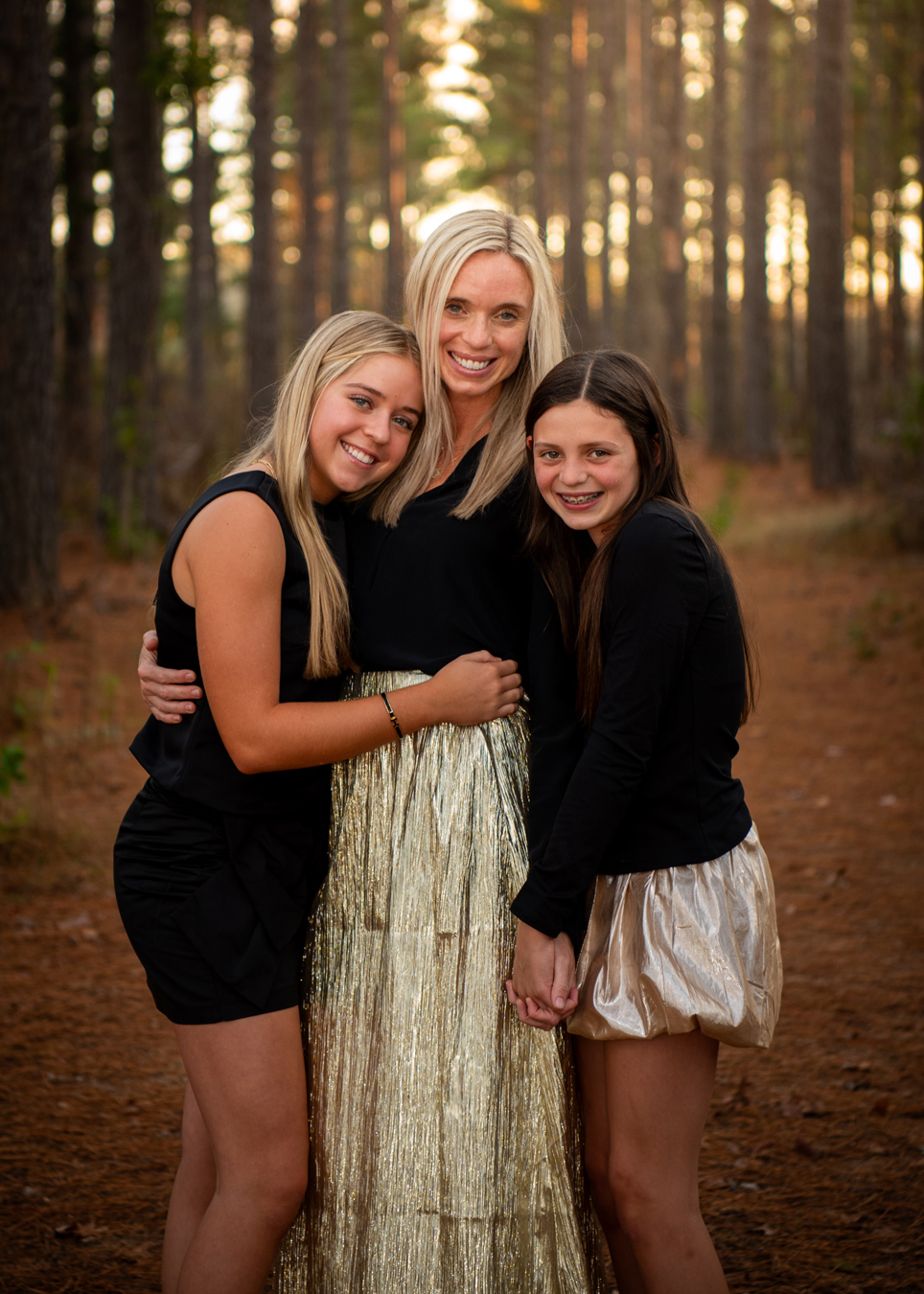 Smiling mother and two daughters in a pine forest. Mother wears a gold skirt; daughters wear black tops and gold bottoms