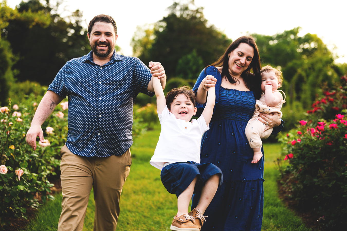 A family of four is walking through a garden, with the father holding his son up by the hands as the boy smiles, and the mother holding a baby in her arms, also smiling. The background features lush greenery and colorful flowers.