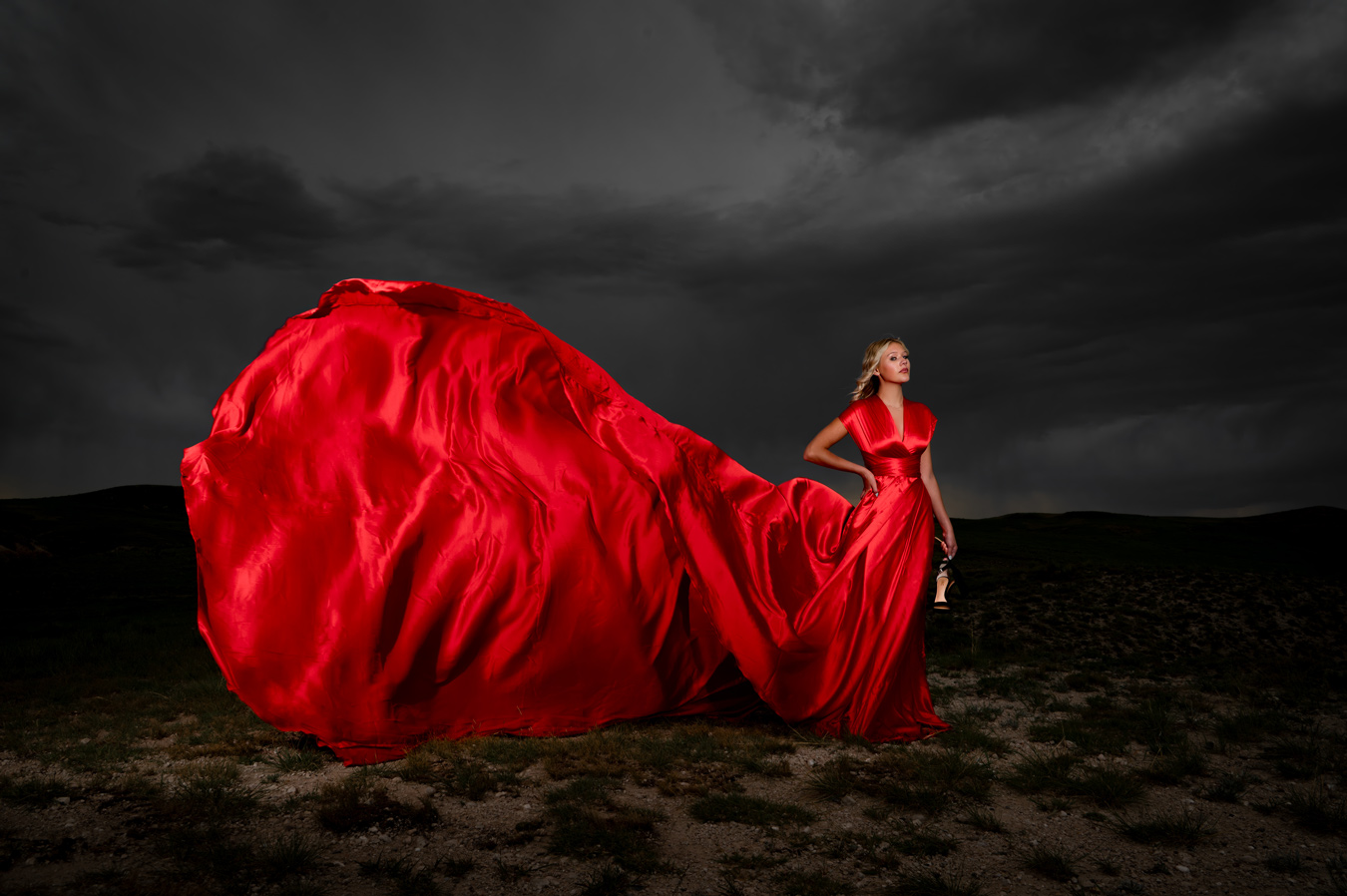 A woman in a vibrant red satin dress stands in a field under a stormy sky, the dress's long train dramatically billowing around her. She holds a pair of shoes in her hand, looking off into the distance. Image by Misty Huss Photography