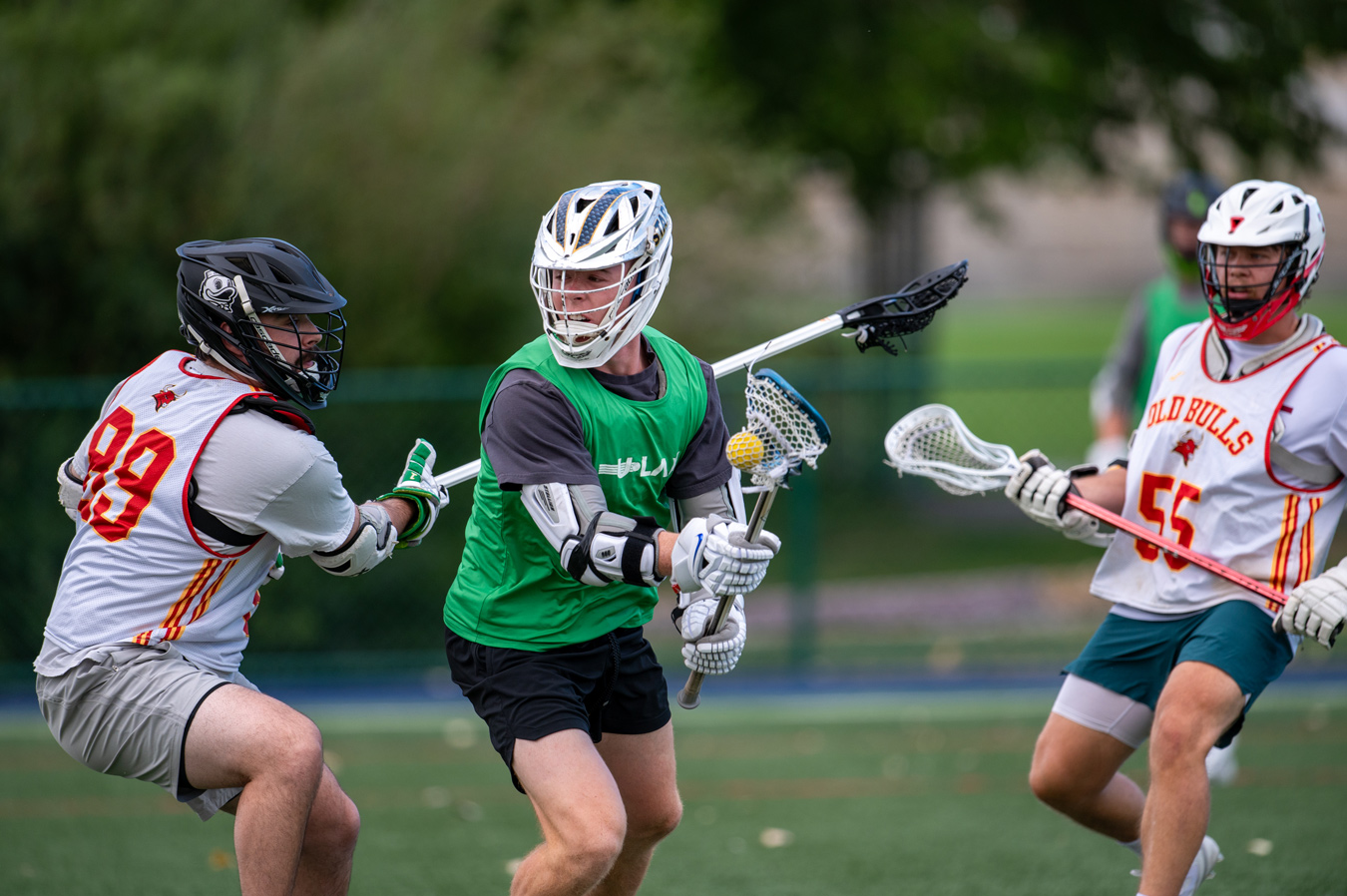 Three lacrosse players in full gear are actively engaged in a game on a green field, with the central player in a green vest holding a lacrosse stick with a ball in its net. The players on either side, wearing team jerseys, are positioned to defend or challenge for possession of the ball. Image by Misty Huss Photography