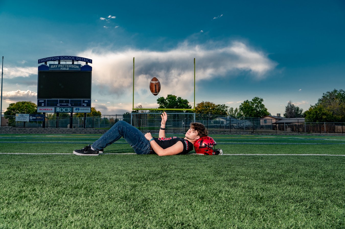 A young man in jeans and a black tank top lies on a football field with his red helmet next to him, reaching up towards a floating football. In the background are goalposts, a scoreboard, and a cloudy sky. Image by Misty Huss Photography