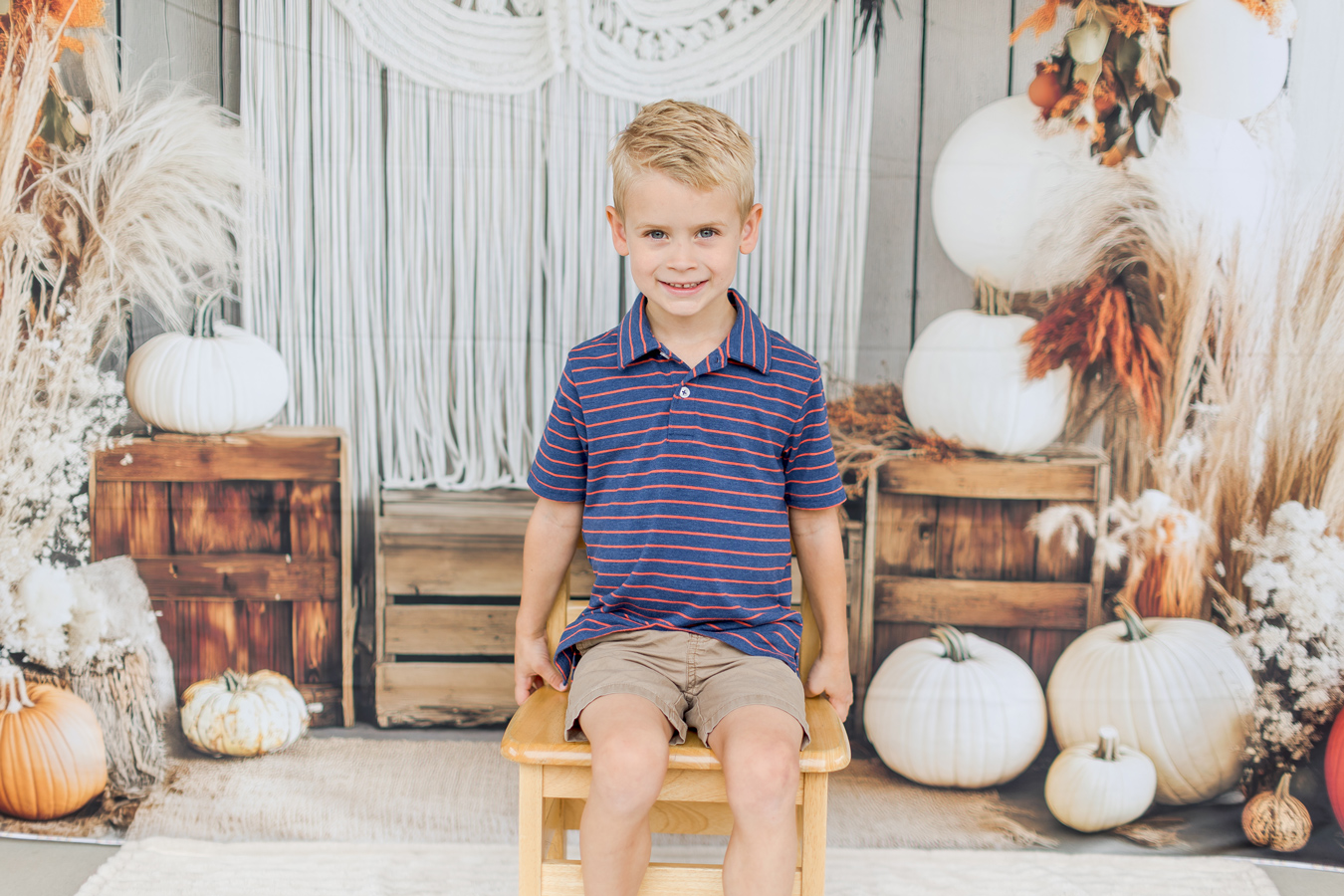 A young boy with blonde hair and a striped shirt sits on a wooden chair, smiling at the camera. The backdrop features white pumpkins, wooden crates, and decorative fall foliage.