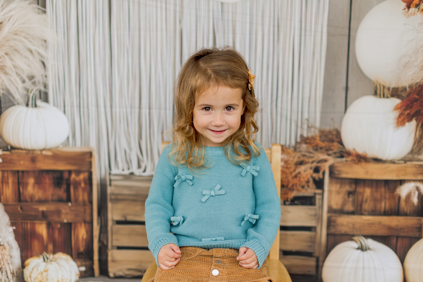 A young girl with curly brown hair and a gold star clip smiles at the camera, wearing a blue sweater with bows and a brown corduroy skirt. The background features white pumpkins, wooden crates, and a white macrame curtain, creating an autumnal scene.