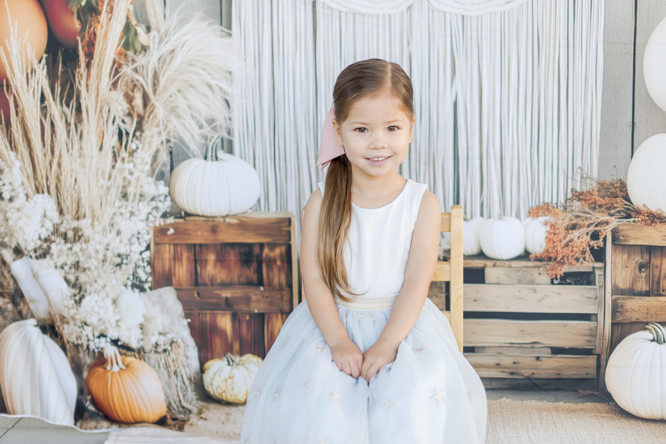 A young girl with a pink bow in her hair sits smiling in a white and blue dress, posed in front of a backdrop of white pumpkins, wooden crates, and dried floral arrangements. The scene has a soft, autumnal aesthetic.