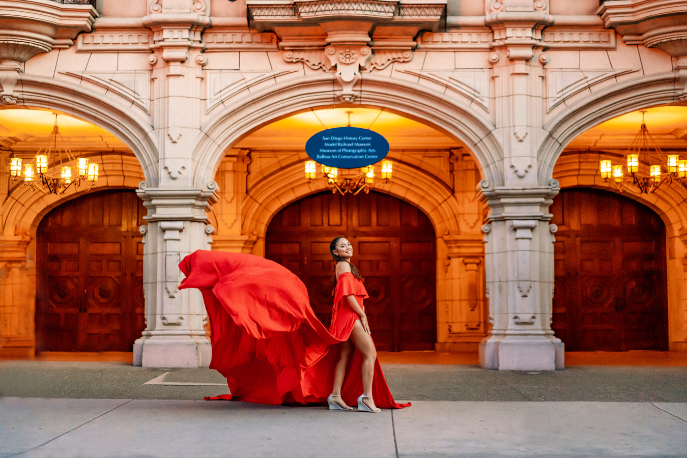 A woman in a red dress stands in front of an ornate building with arched doorways and chandeliers. The dress has a long train that is blowing in the wind. Photo credit: Czarina Luna