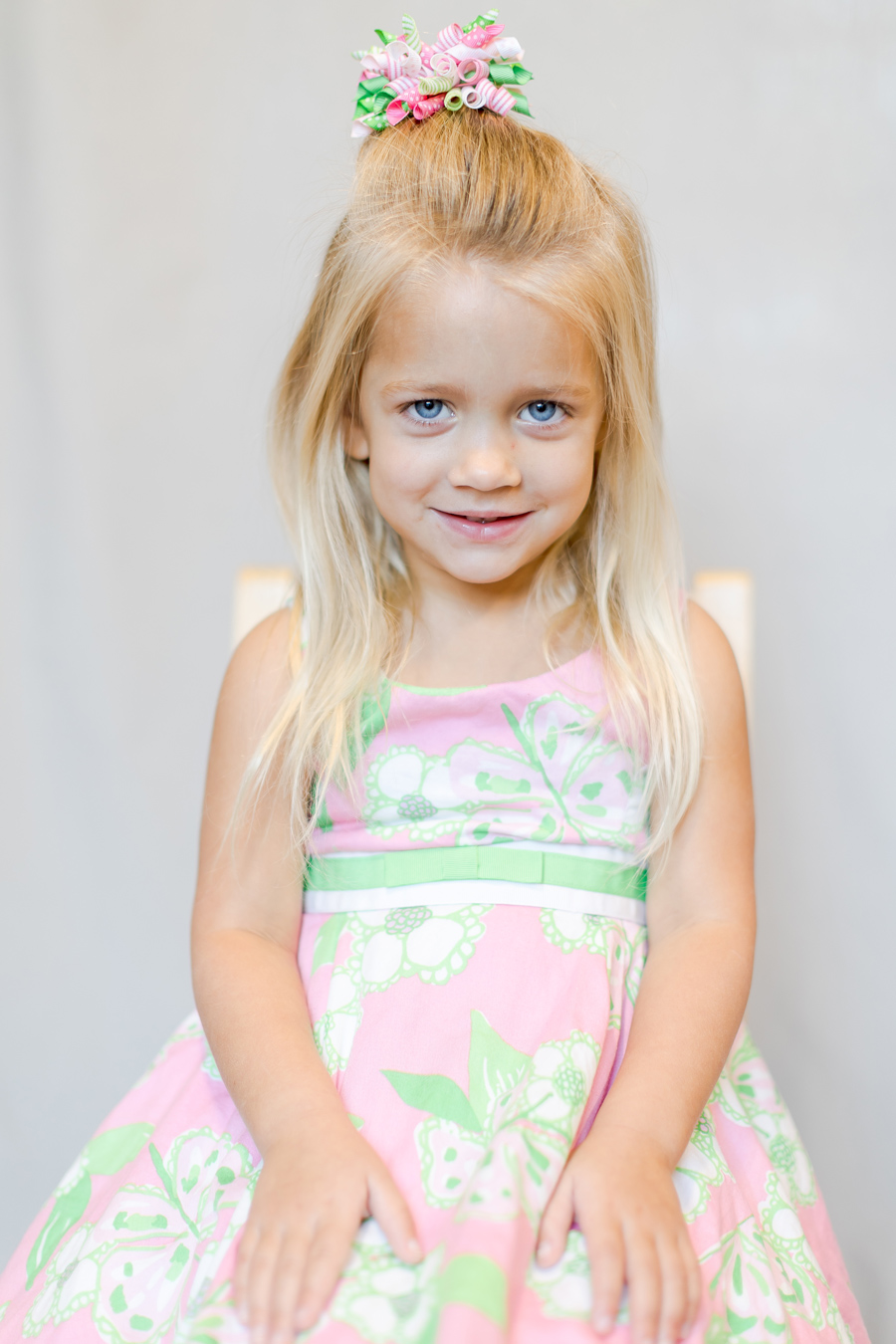 A young girl with blonde hair and bright blue eyes smiles at the camera, wearing a pink and green floral dress and a matching ribbon hair accessory. She is seated against a neutral background.