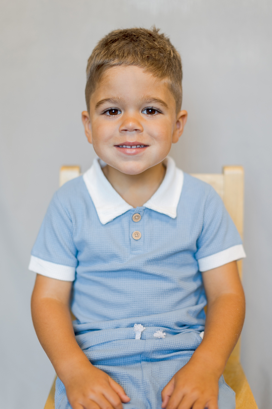A young boy with brown hair and eyes smiles at the camera, wearing a light blue waffle-textured polo shirt with a white collar and matching shorts. He is sitting on a wooden chair against a neutral background.