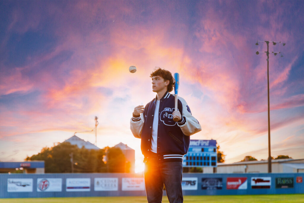 A young man in a letterman jacket stands on a baseball field with a bat over his shoulder, looking up at a baseball floating in the air. The background features a vibrant sunset sky and a baseball field with stadium lights. Photo credit: Dustin Armstrong