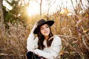 A young woman with long brown hair sits in a field of tall, dry grass, wearing a cream-colored sweater and a black hat, looking off to the side with a gentle smile. The sunlight filters through the trees in the background, creating a warm, golden glow. Photo credit: LKW Photography