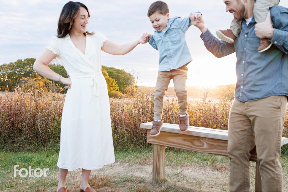 A family is enjoying time outdoors, with a smiling mother in a white dress holding hands with her young son as he jumps off a wooden bench, while the father stands nearby with another child on his shoulders. The scene is set in a field with trees in the background, bathed in the warm glow of the setting sun.