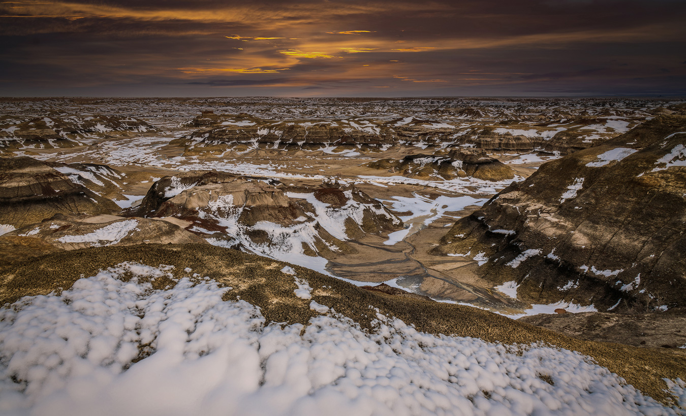 A wide, scenic view shows a vast, arid landscape of layered rock formations dusted with snow under a dramatic, cloudy sunset sky. The foreground features a snow-covered ridge, leading to a panorama of canyons and mesas stretching towards the horizon at the Bisti Badlands.