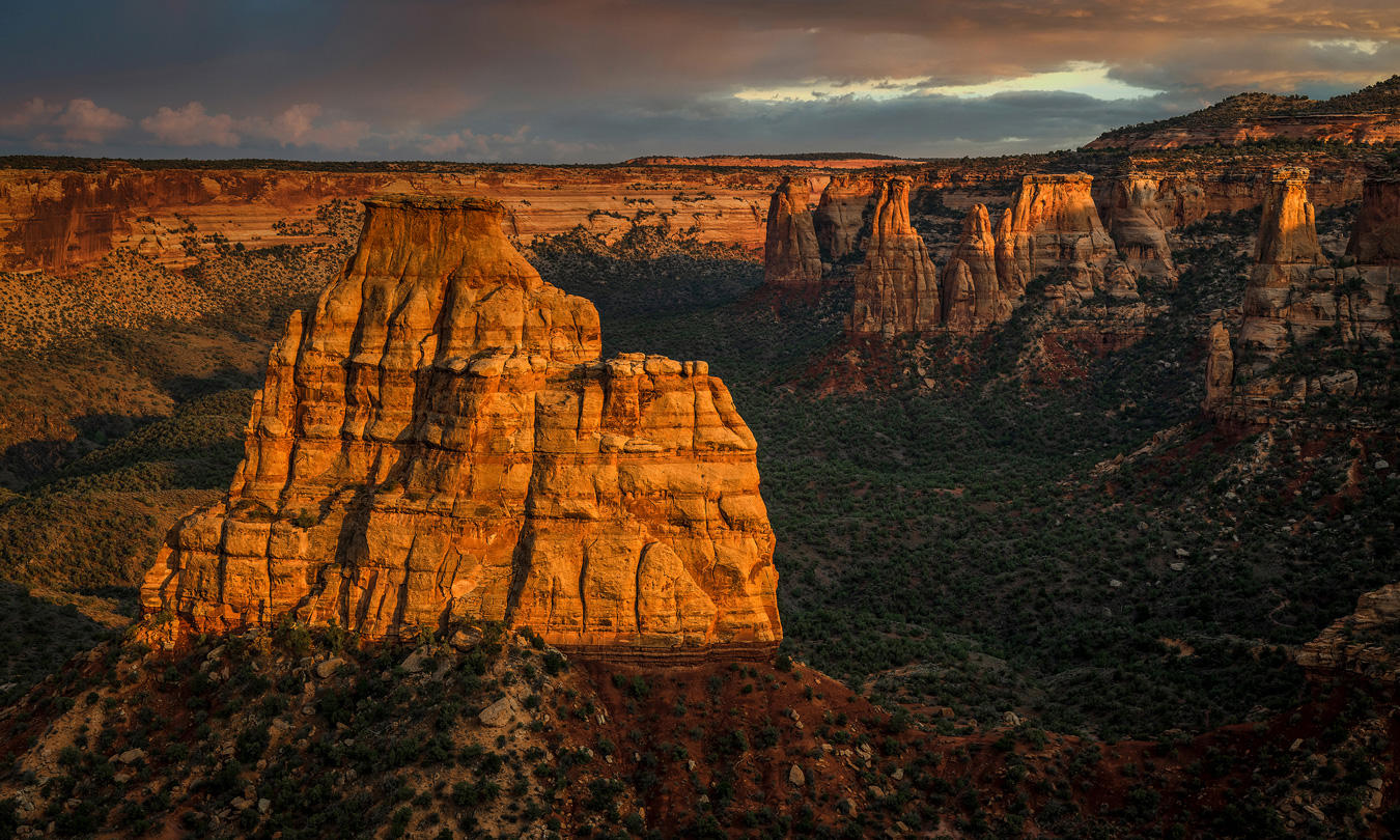 A scenic landscape features a large, layered sandstone formation bathed in golden light, with a canyon and other rock formations visible in the background under a dramatic sky. The foreground is covered in green vegetation, contrasting with the warm tones of the rock.