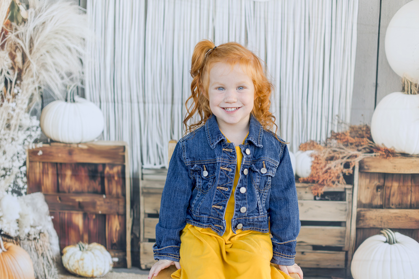 A young girl with red hair pulled into a ponytail smiles at the camera, wearing a yellow dress and denim jacket. She is sitting in front of a backdrop of white pumpkins, wooden crates, and a macrame curtain.