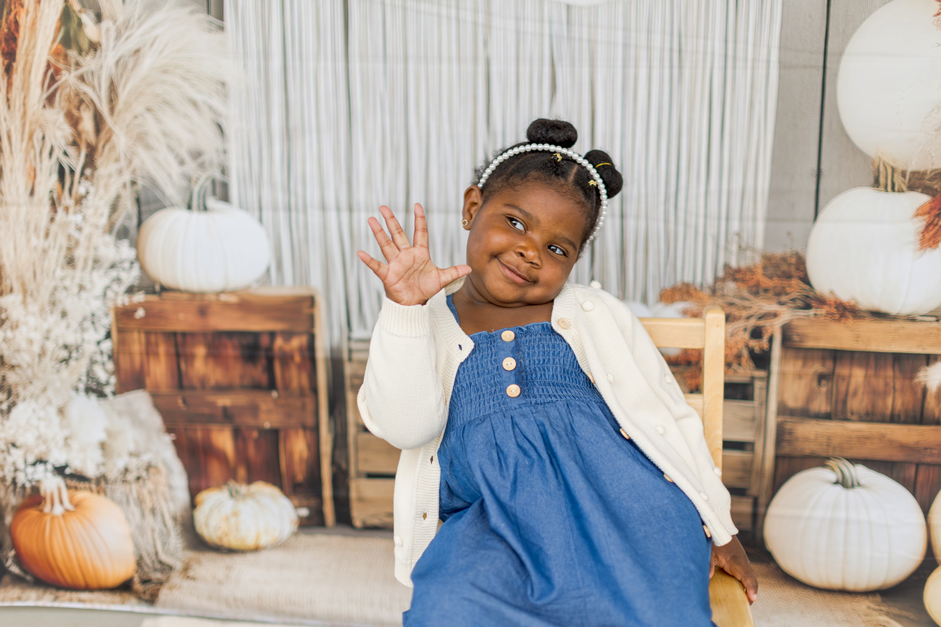 A young Black girl with her hair in buns and a pearl headband smiles and waves, wearing a blue dress and cream cardigan. She is sitting in a chair against a backdrop of pumpkins, wooden crates, and white curtains.