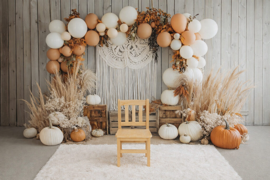 A light wooden chair sits on a white rug in front of a backdrop decorated with white and tan balloons, pampas grass, white pumpkins, and a macrame wall hanging. The scene is set against a light gray wooden wall, creating a cozy and autumnal atmosphere.