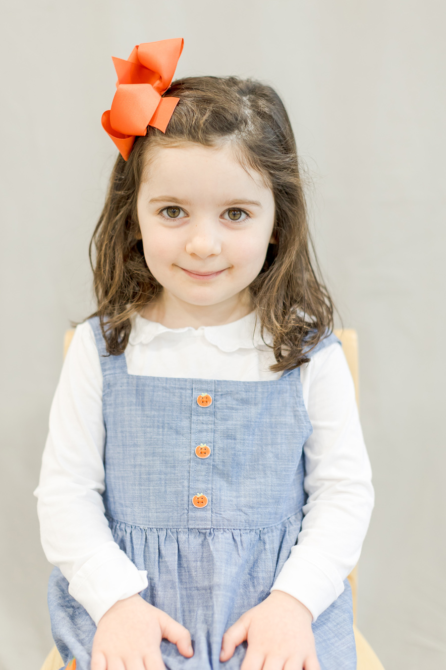 A young girl with brown hair and eyes smiles at the camera, wearing a blue dress with pumpkin-shaped buttons and a large orange bow in her hair. She is sitting on a chair against a neutral background.