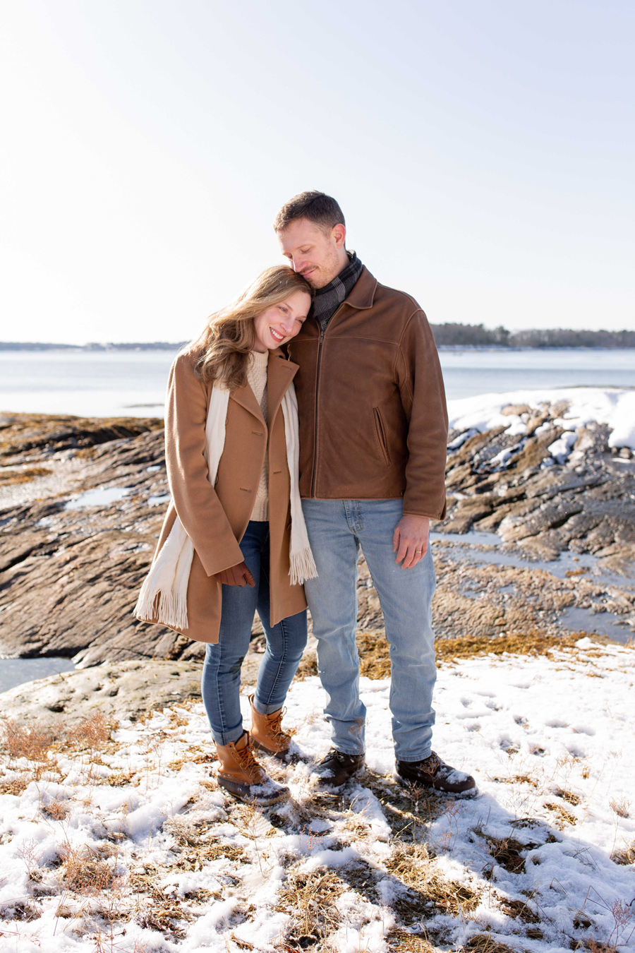 A smiling woman rests her head on a man's shoulder as they stand together on a snowy, rocky beach. The woman wears a tan coat and boots, while the man wears a brown leather jacket and scarf. Photo by Wild Orchard Studios