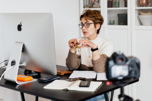 A woman with short brown hair and glasses sits at a desk with an Apple computer, holding a small gold object, likely recording a video with a DSLR camera positioned in the foreground. The desk also contains a notebook, phone, calculator, and other items, with a white cabinet visible in the background.