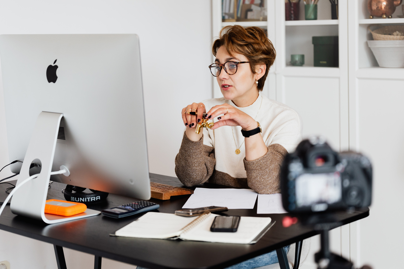 A woman with short brown hair and glasses sits at a desk with an Apple computer, holding a small gold object, likely recording a video with a DSLR camera positioned in the foreground. The desk also contains a notebook, phone, calculator, and other items, with a white cabinet visible in the background.